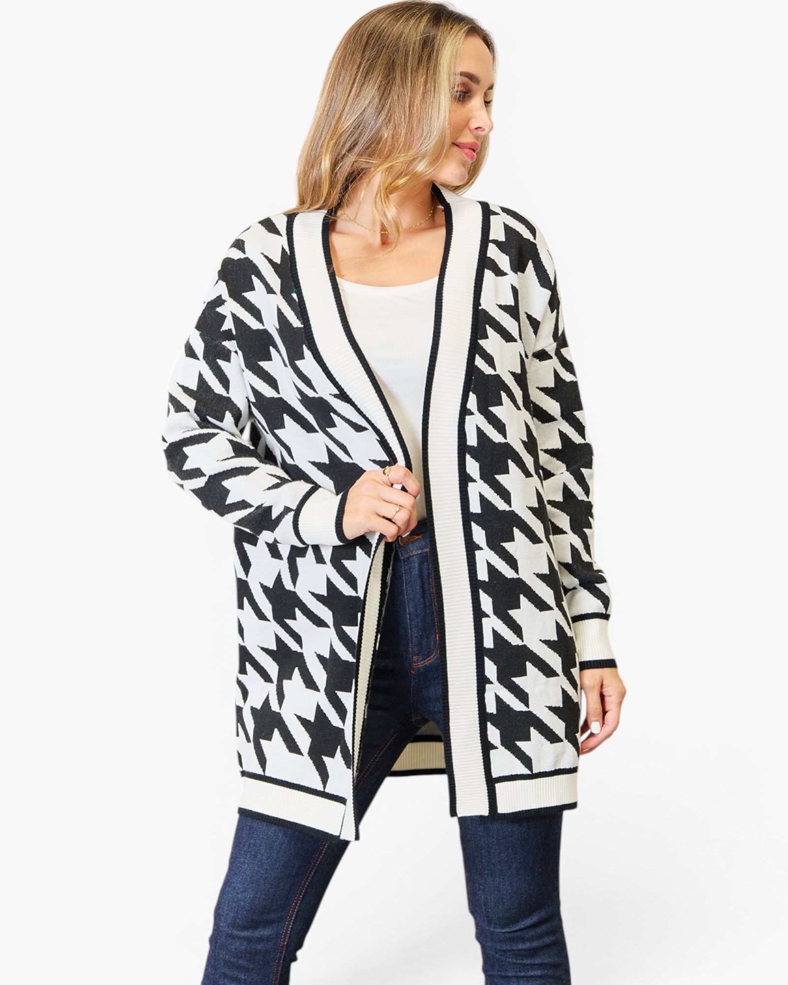 A woman modeling a black and white houndstooth pattern cardigan with black trim over a white top and blue jeans, against a plain white background.