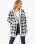 A woman modeling a black and white houndstooth pattern cardigan with black trim over a white top and blue jeans, against a plain white background.
