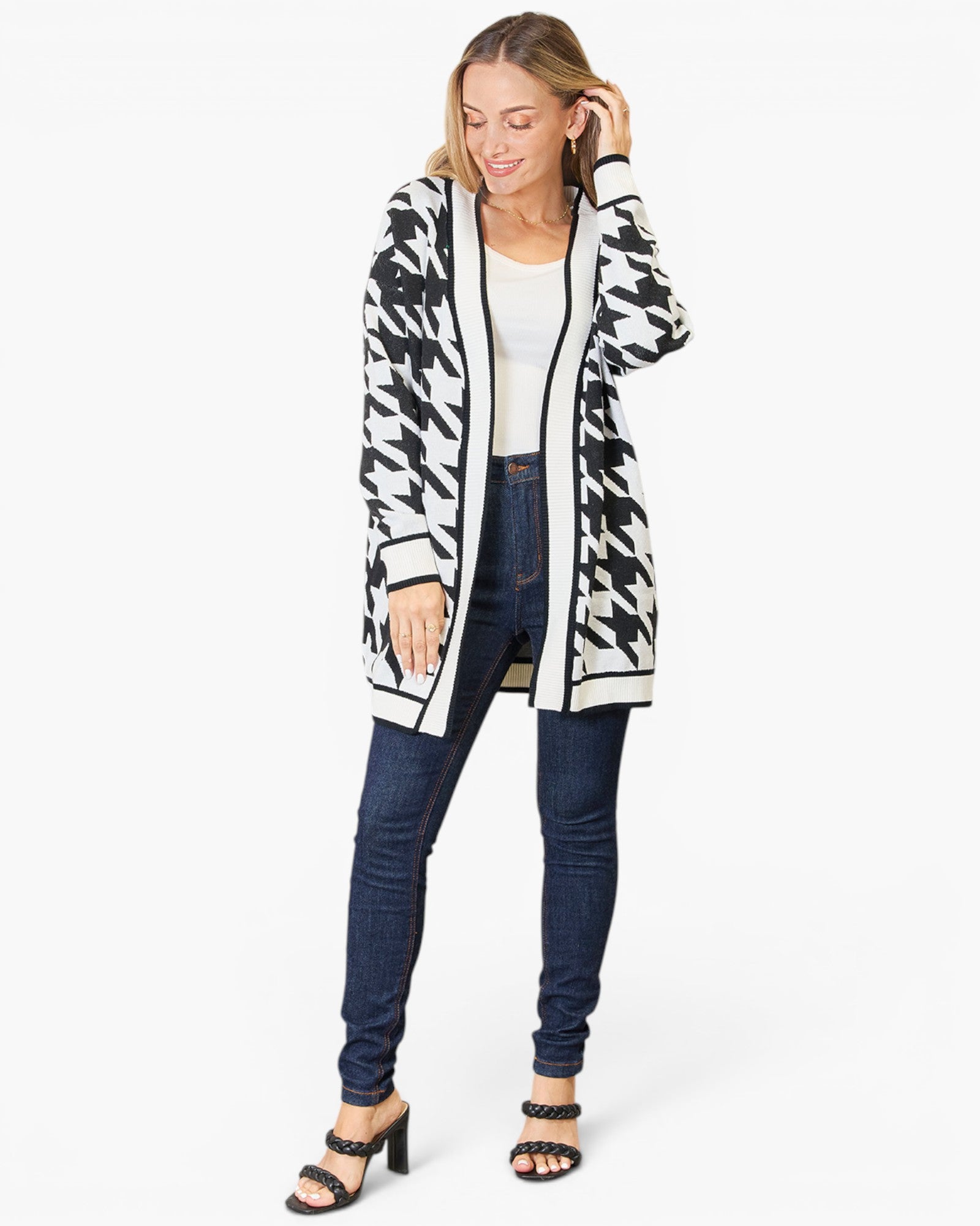 A woman modeling a black and white houndstooth open-front cardigan over a white top and dark blue skinny jeans, standing against a light gray studio background.