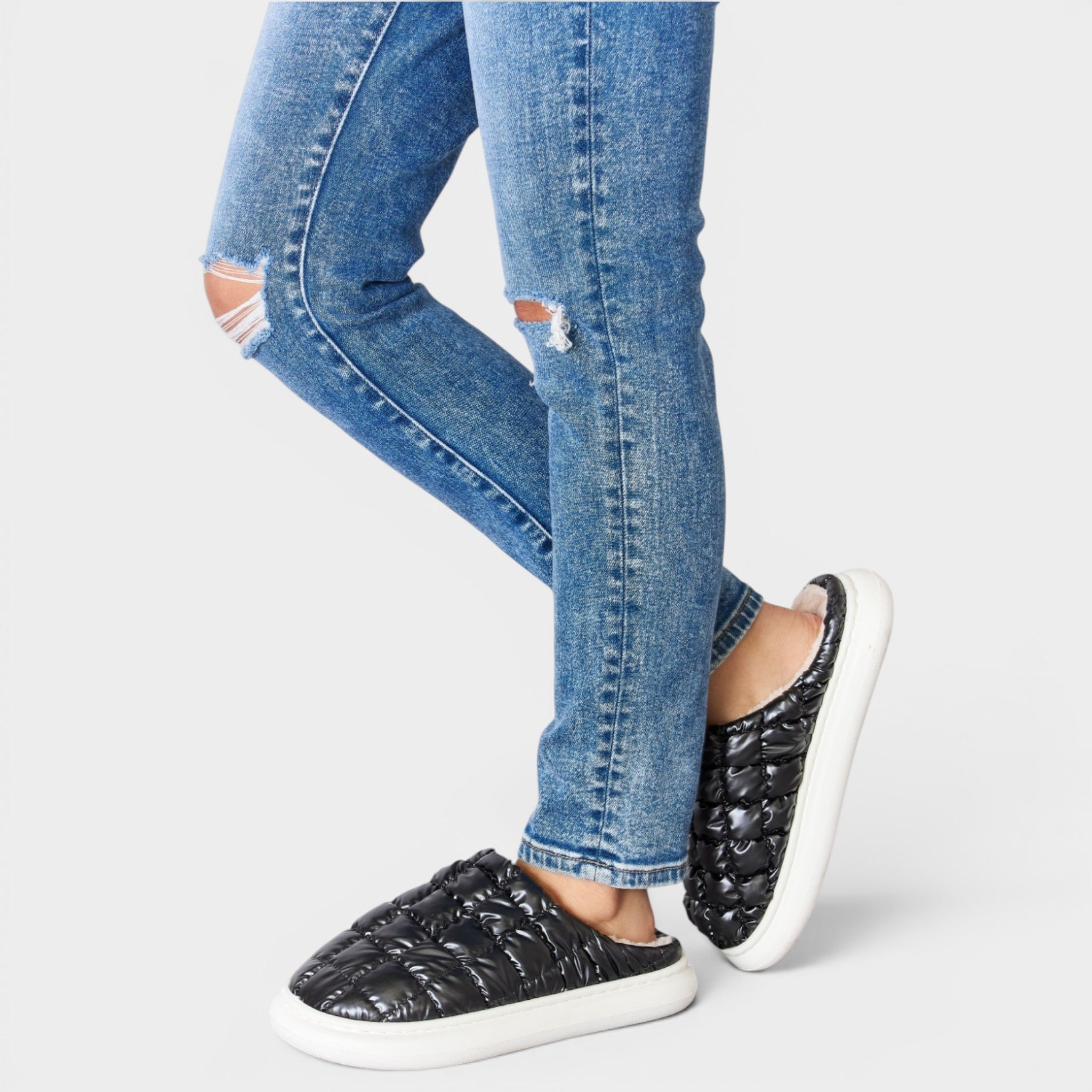 Person wearing blue jeans and black slip-on shoes with white soles on a light gray background