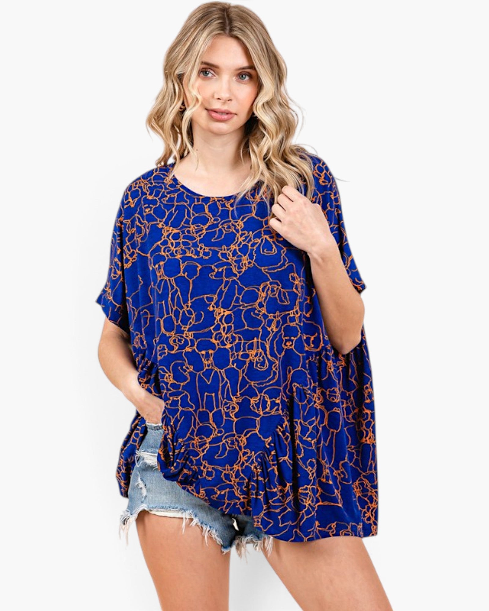 A woman modeling a loose, short-sleeve blue top with an orange abstract line pattern, paired with denim shorts, against a plain white studio background.