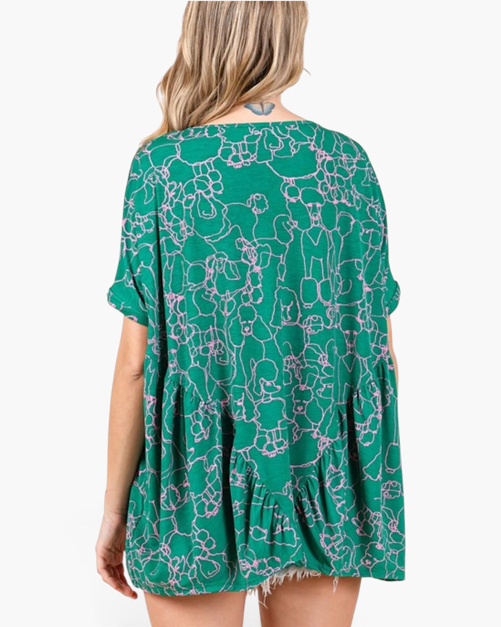 A woman shown from the back wearing a green short-sleeve tunic dress with a white line-art dog pattern and a ruffled hem, set against a plain white background.