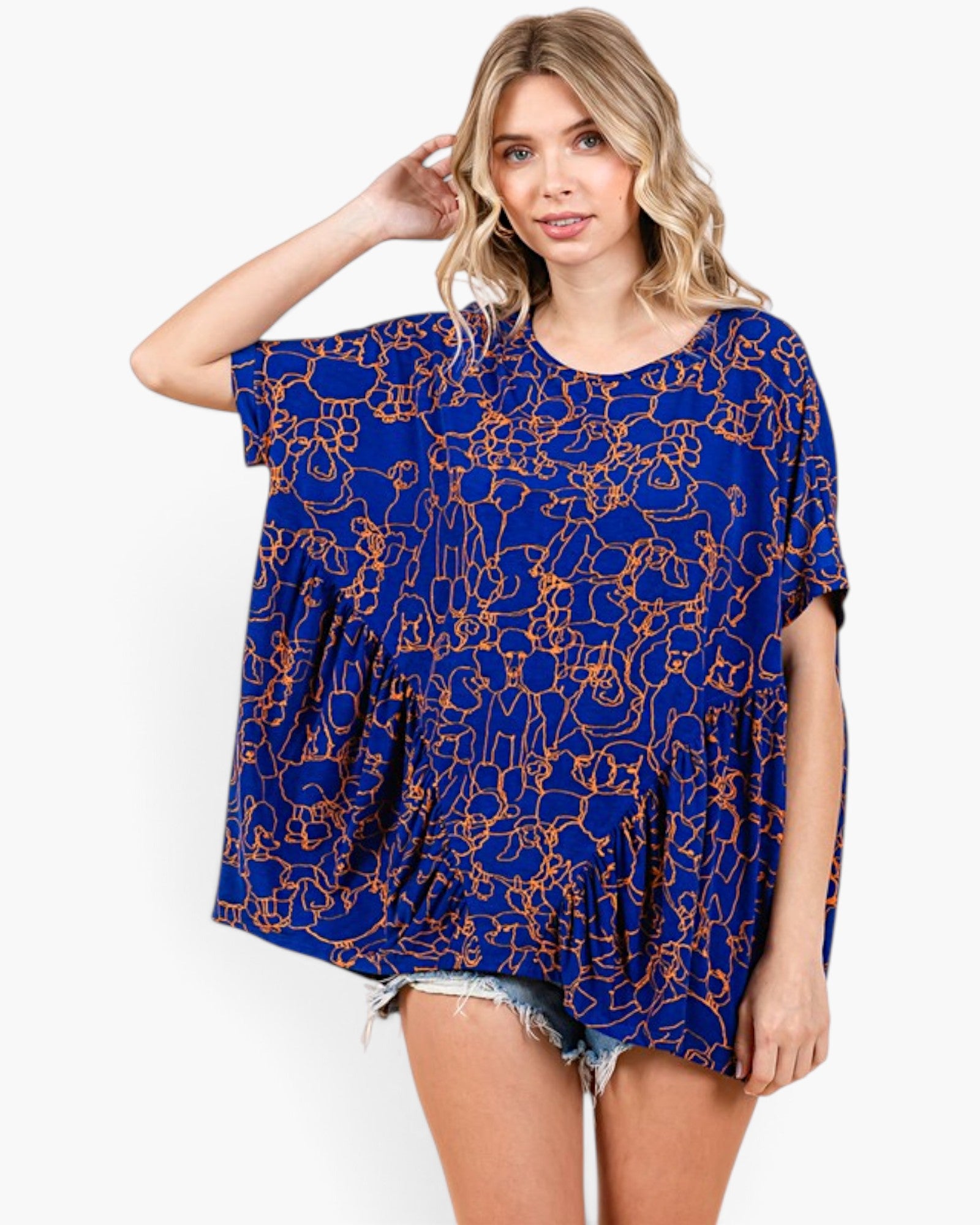 A woman modeling a loose, short-sleeve blue top with an orange abstract line pattern, paired with denim shorts, against a plain white studio background.