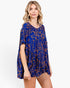 A woman modeling a loose short-sleeve tunic dress with a vibrant blue base and an orange abstract line pattern, set against a plain white studio background.