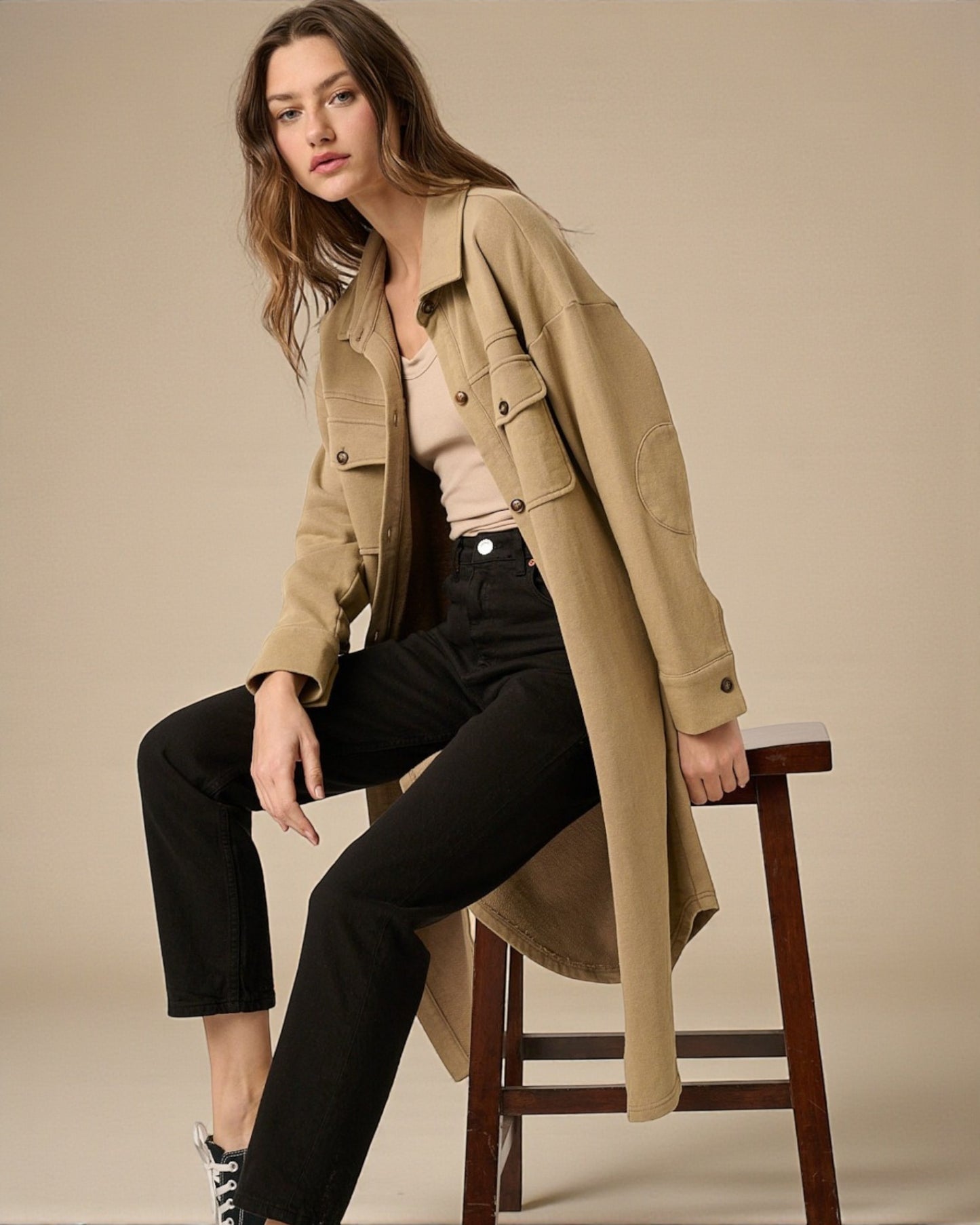 Woman wearing a beige trench coat sitting on a chair against a plain background