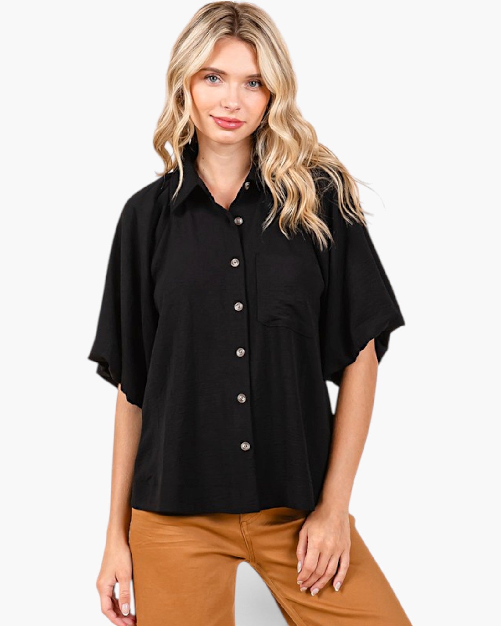A woman modeling a black short-sleeve button-up shirt with a relaxed, oversized fit and brown pants, set against a plain light gray background.