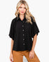 A woman modeling a black short-sleeve button-up shirt with a relaxed, oversized fit and brown pants, set against a plain light gray background.