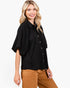 A woman modeling a black short-sleeve button-up blouse with a relaxed fit and brown pants, photographed against a clean white studio background.