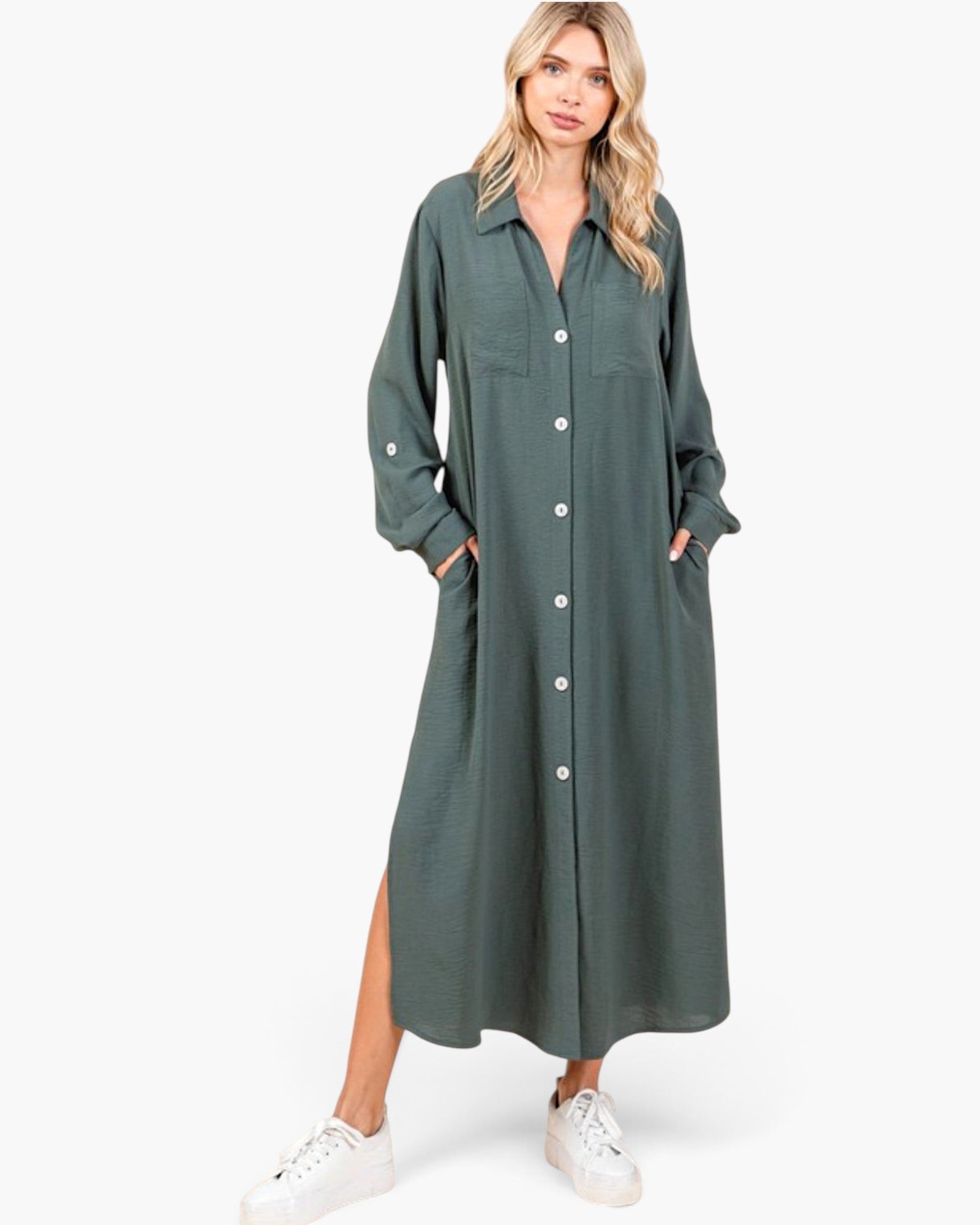 A woman modeling a long-sleeve, button-front midi shirt dress in a muted sage green color, styled with white sneakers against a plain white studio background.