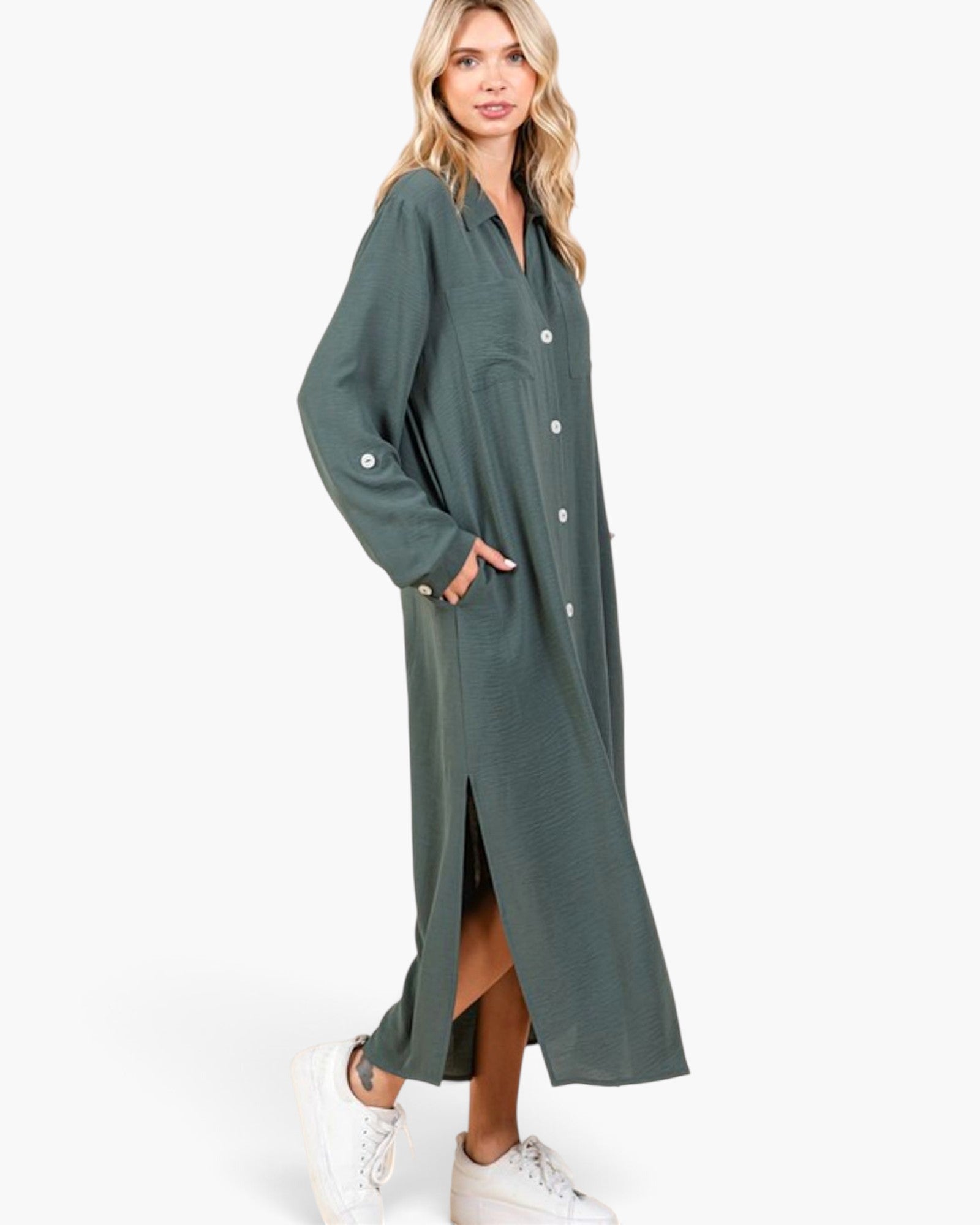 A woman modeling a long-sleeve, button-front midi dress in a muted sage green color, styled with white sneakers against a plain white studio background.