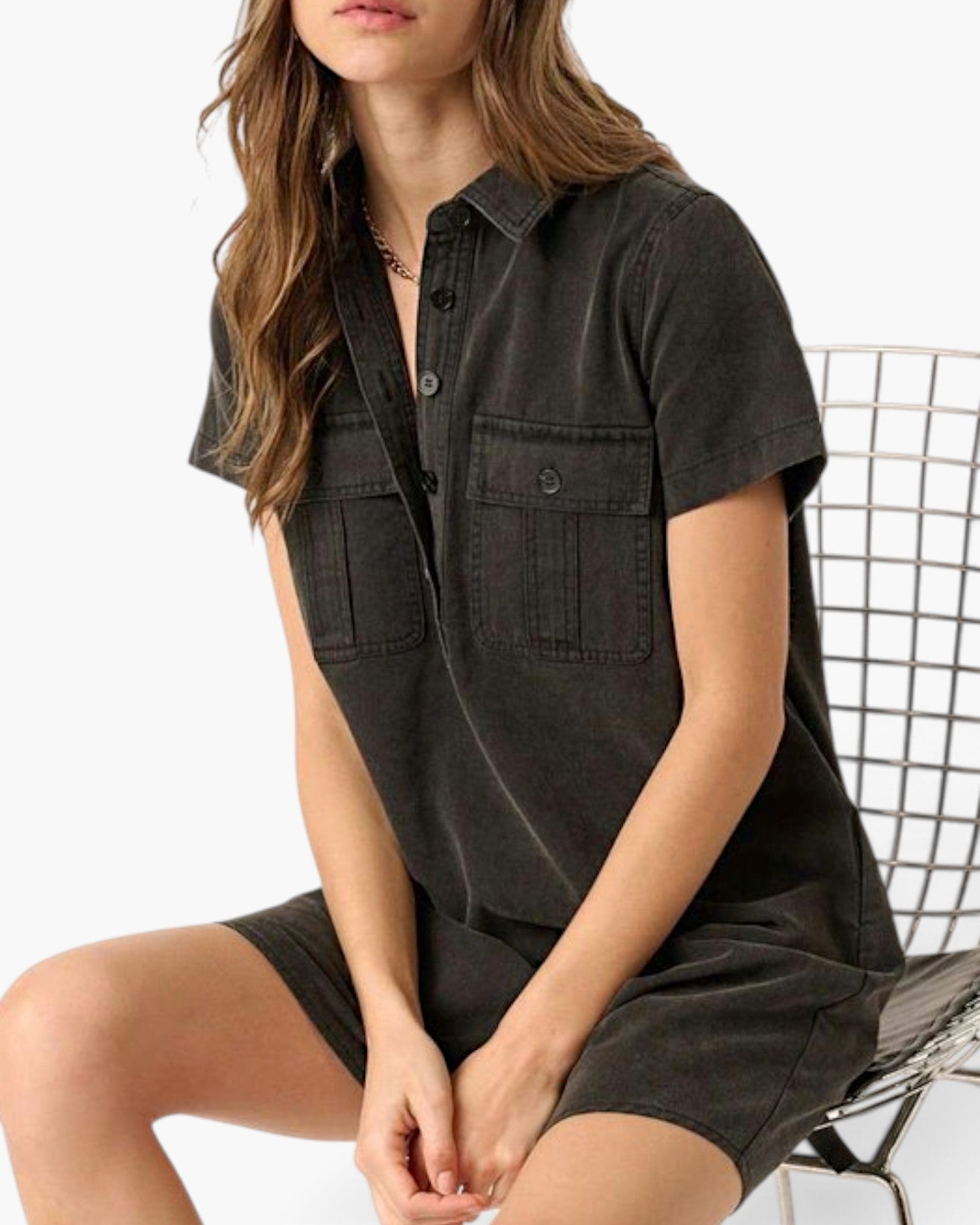 A woman wearing a dark charcoal gray short-sleeve button-up shirt dress, seated on a wire chair against a white background.