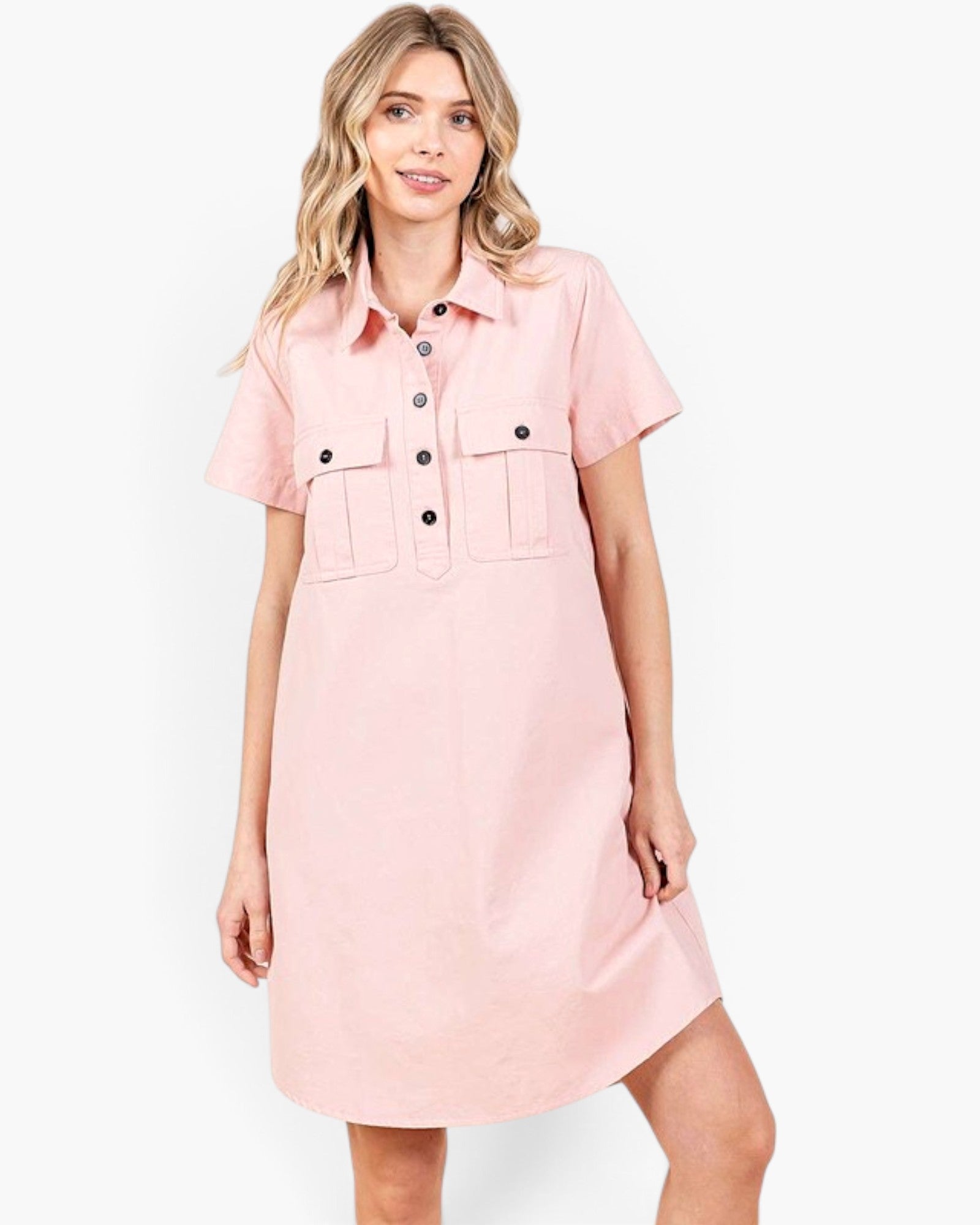 A woman modeling a light pink short-sleeve shirt dress with a button-down front and two chest flap pockets, photographed against a plain light gray studio background.