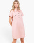 A woman modeling a light pink short-sleeve shirt dress with a button-down front and two chest pockets, photographed against a clean white studio background.