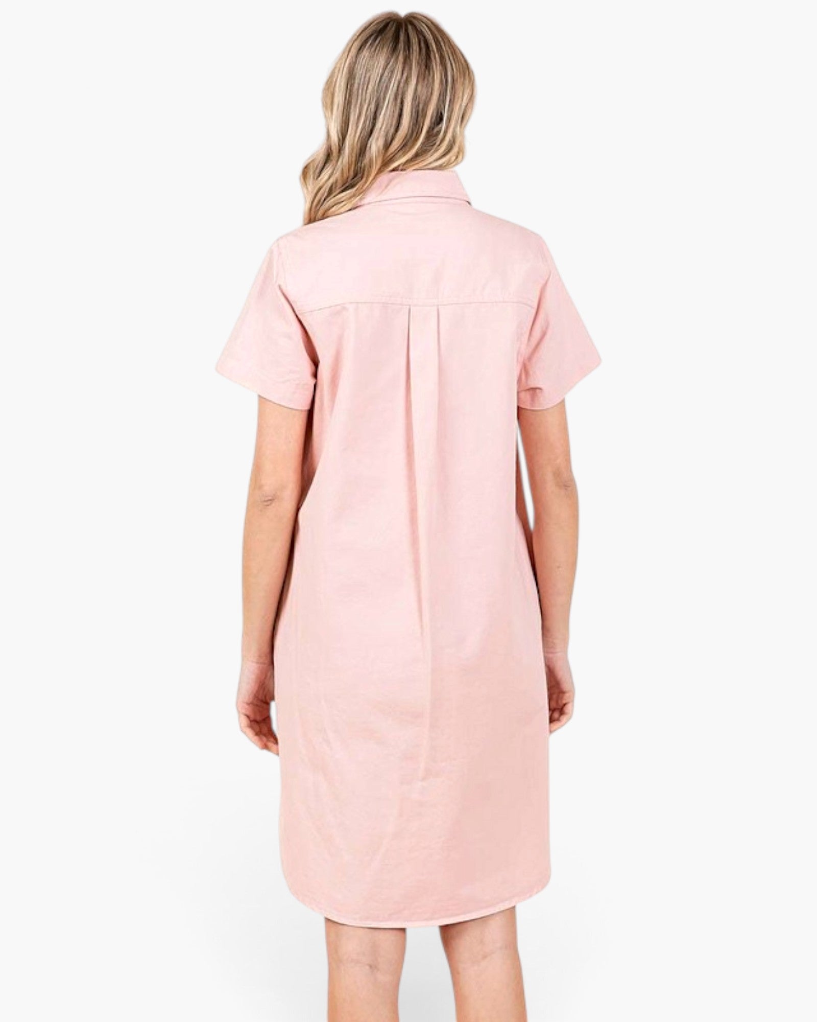 A woman viewed from the back wearing a short-sleeve pink shirt dress with a button-down style and a central back pleat, set against a plain white background.