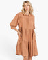 A woman modeling a tiered, long-sleeve button-up shirt dress in a warm terracotta or rust color, set against a plain white studio background.
