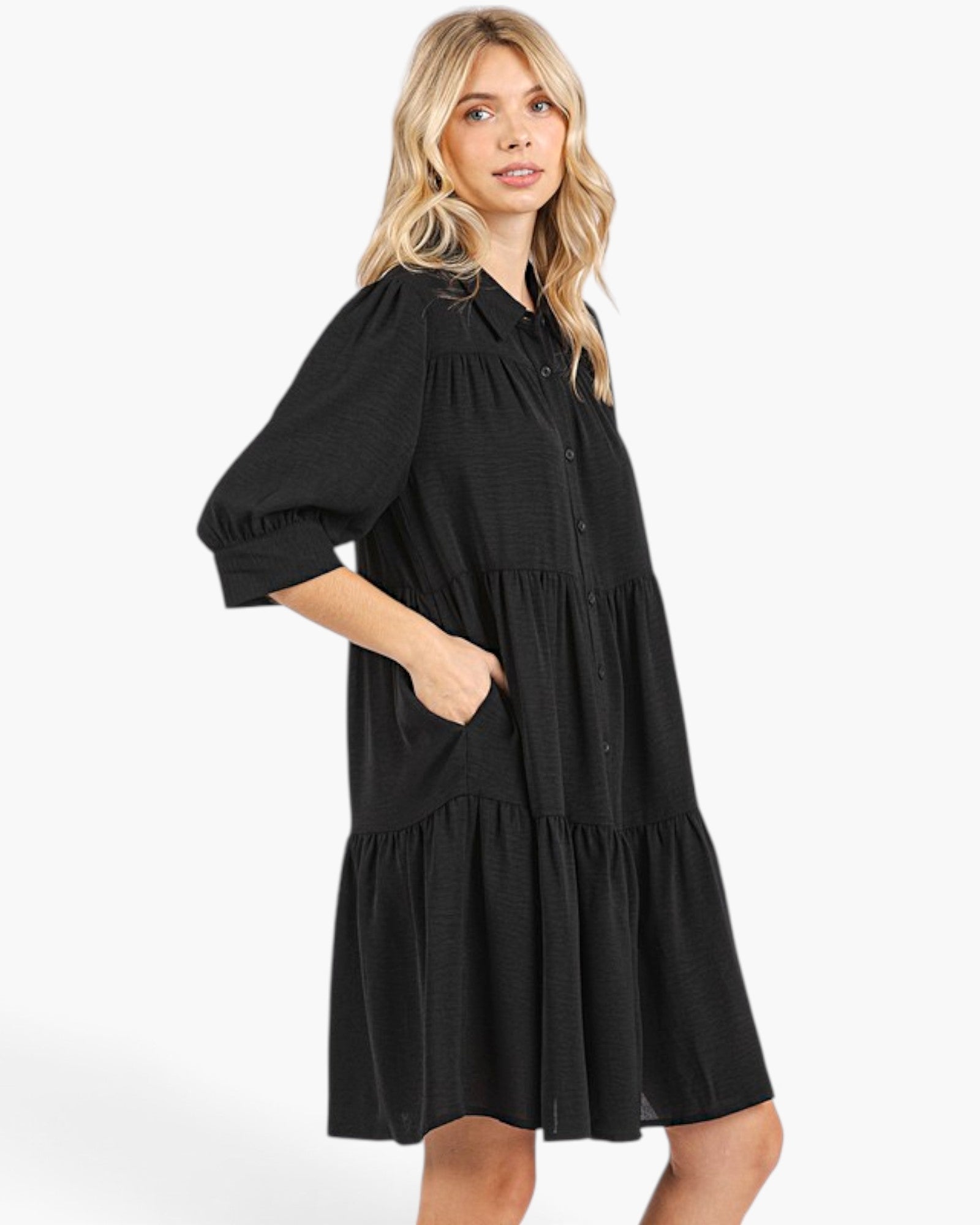 A woman modeling a black tiered midi dress with puff sleeves, a button-down front, and side pockets, photographed against a clean white studio background.