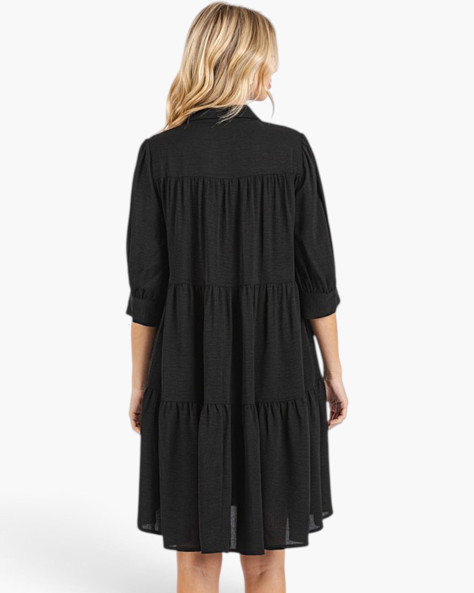 A woman viewed from the back wearing a black tiered midi dress with three-quarter sleeves and a gathered neckline, photographed against a plain light gray studio background.