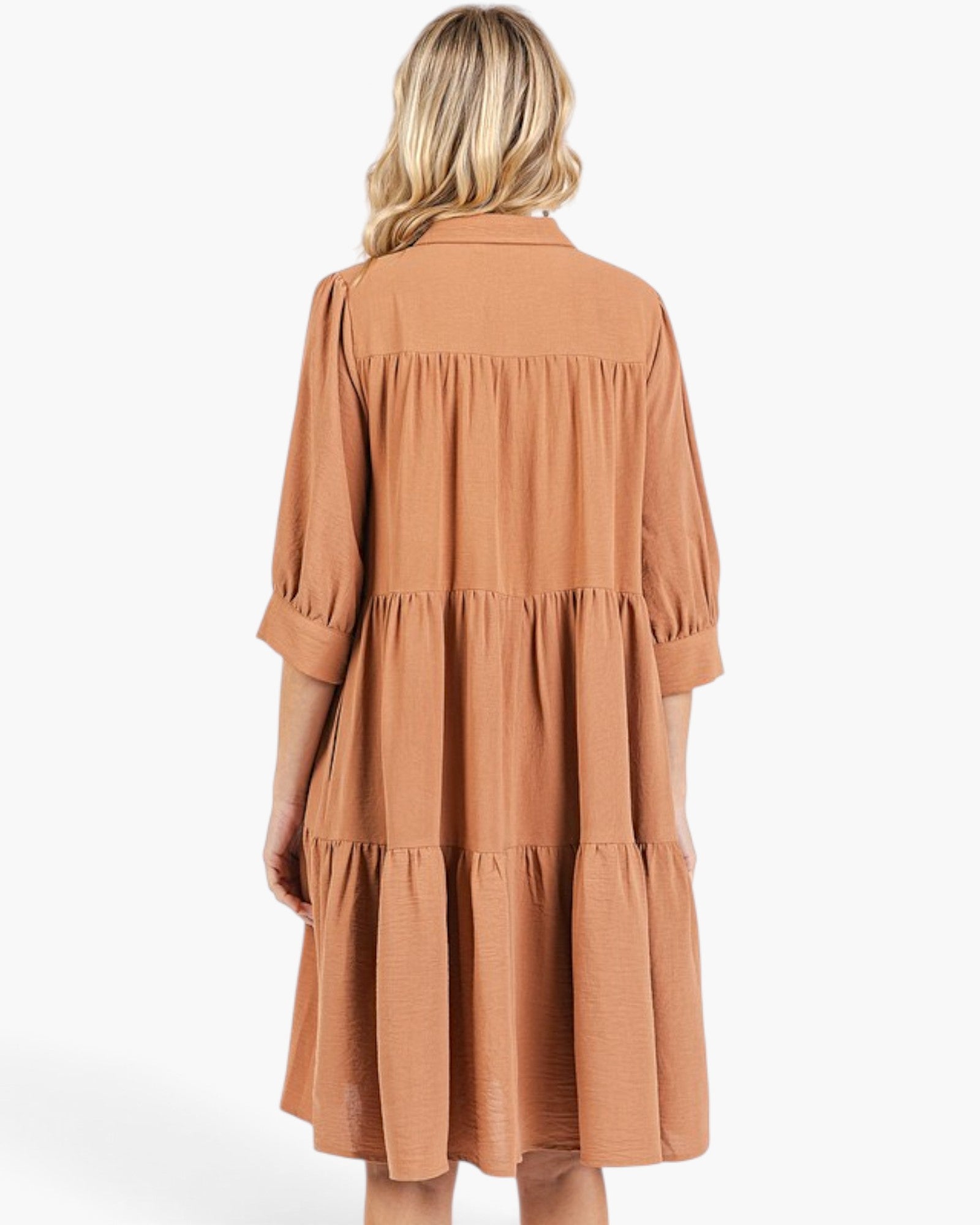 A woman viewed from the back wearing a tiered, three-quarter sleeve dress in a warm terracotta or rust color, set against a plain white studio background.