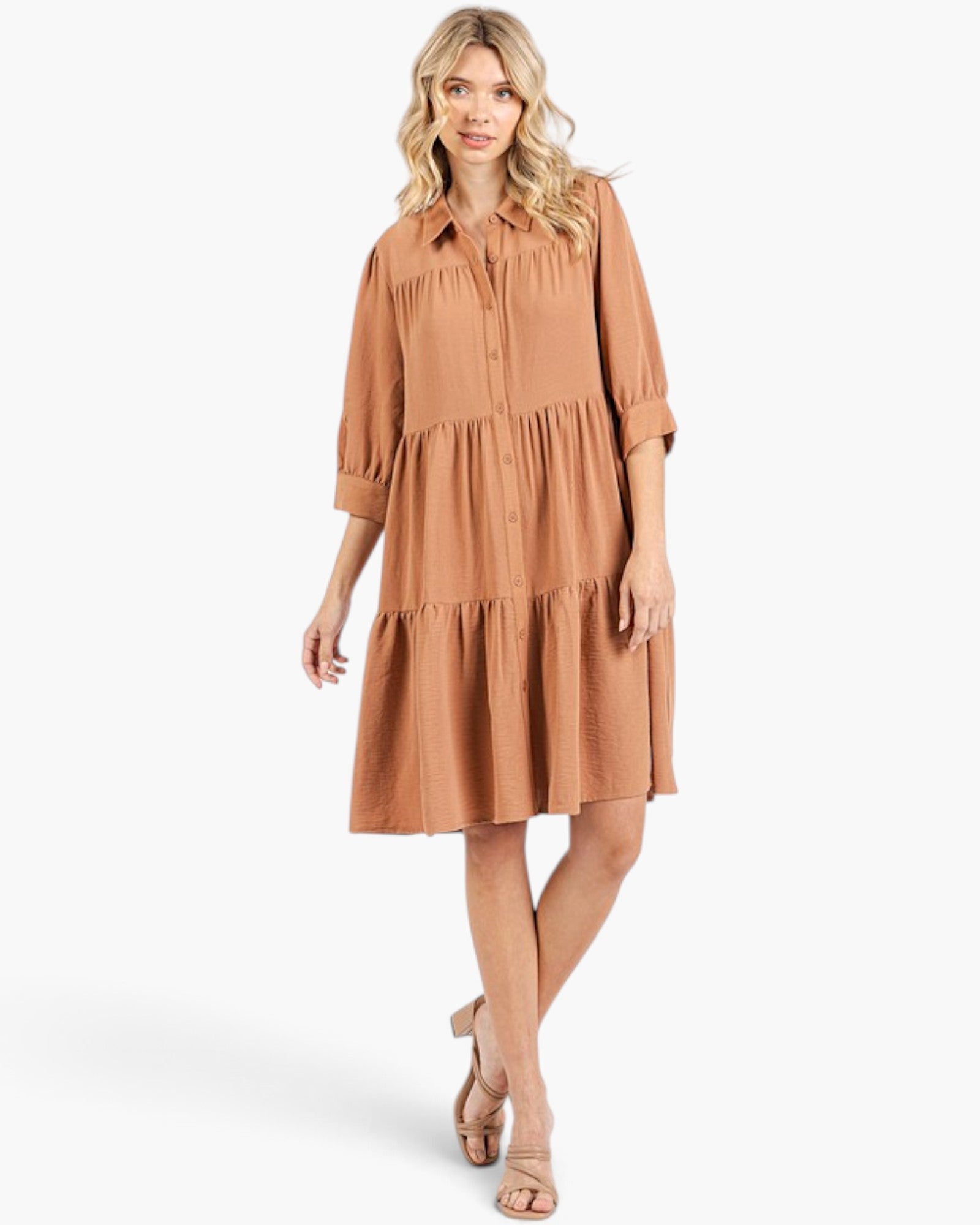 A woman modeling a tiered midi shirt dress in a warm camel brown color, paired with beige heeled sandals, against a plain white studio background.