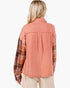 A woman shown from the back wearing a long-sleeve collared shirt with a washed coral-pink body and plaid sleeves in brown, orange, and beige tones, paired with light beige pants against a plain white background.
