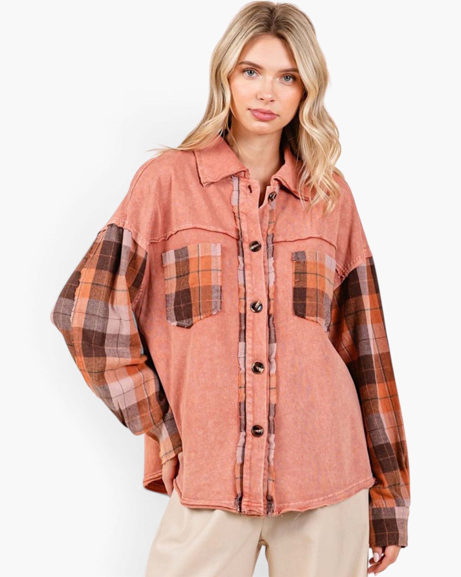 A woman wearing a color-blocked button-up shirt jacket with a dusty rose body and brown, orange, and beige plaid sleeves and chest pockets, paired with beige pants against a white studio background.