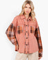 A woman wearing a color-blocked button-up shirt jacket with a dusty rose body and brown, orange, and beige plaid sleeves and chest pockets, paired with beige pants against a white studio background.