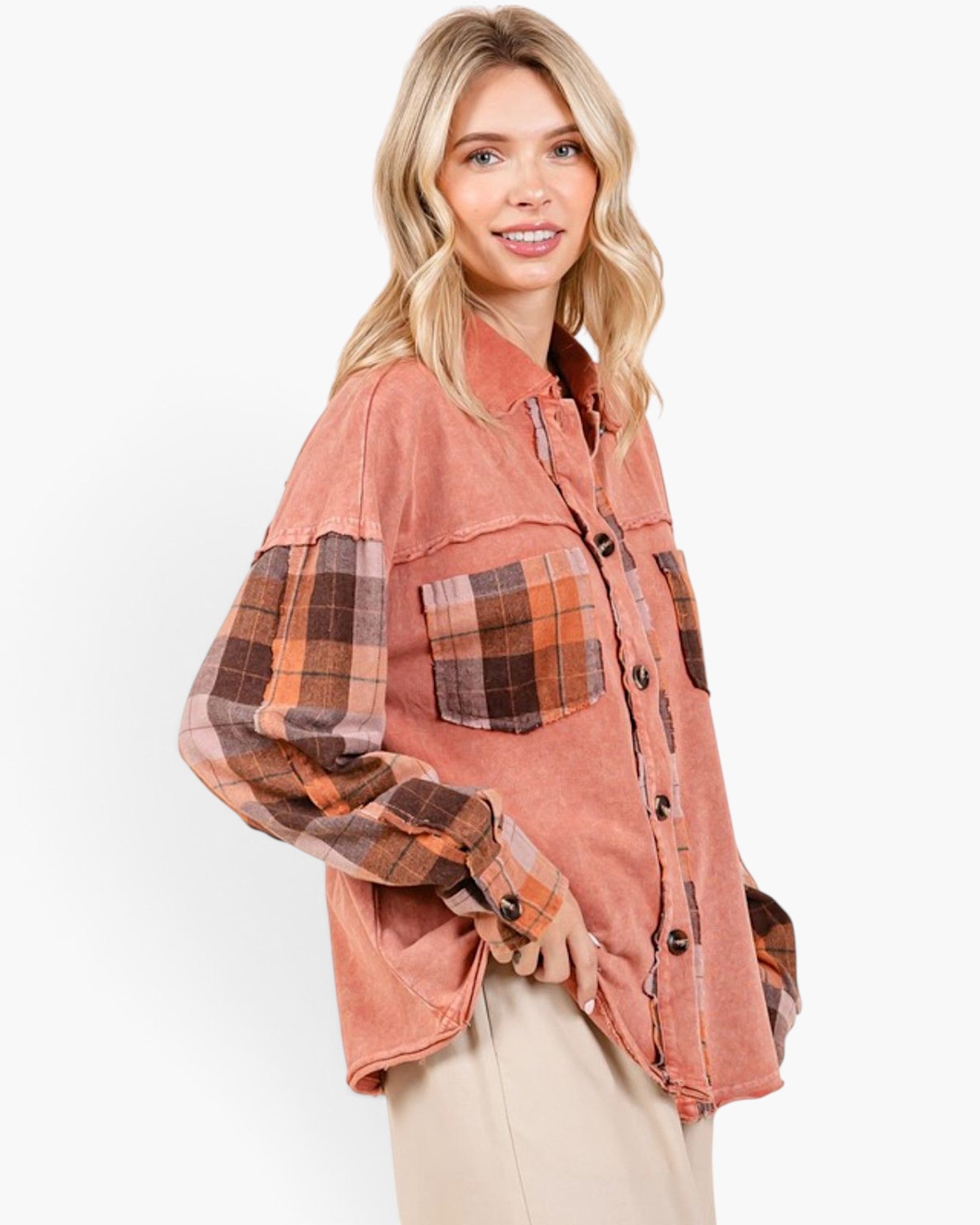 A woman modeling a cropped, oversized button-up shirt with a washed terracotta pink body and contrasting plaid sleeves and chest pockets in brown, orange, and beige tones, paired with beige pants against a plain white background.