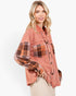 A woman modeling a cropped, oversized button-up shirt with a washed terracotta pink body and contrasting plaid sleeves and chest pockets in brown, orange, and beige tones, paired with beige pants against a plain white background.