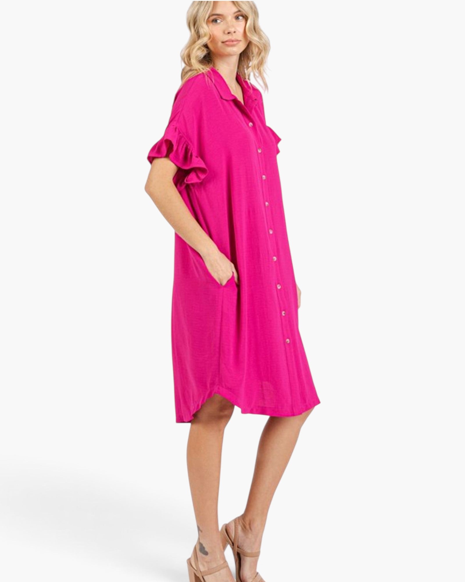 A woman modeling a bright fuchsia pink short-sleeve button-up shirt dress with ruffled sleeves and side pockets, paired with beige high-heeled sandals against a clean white studio background.