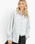 A woman wearing a light blue and white vertically striped button-down shirt with black pants, photographed against a clean white studio background.
