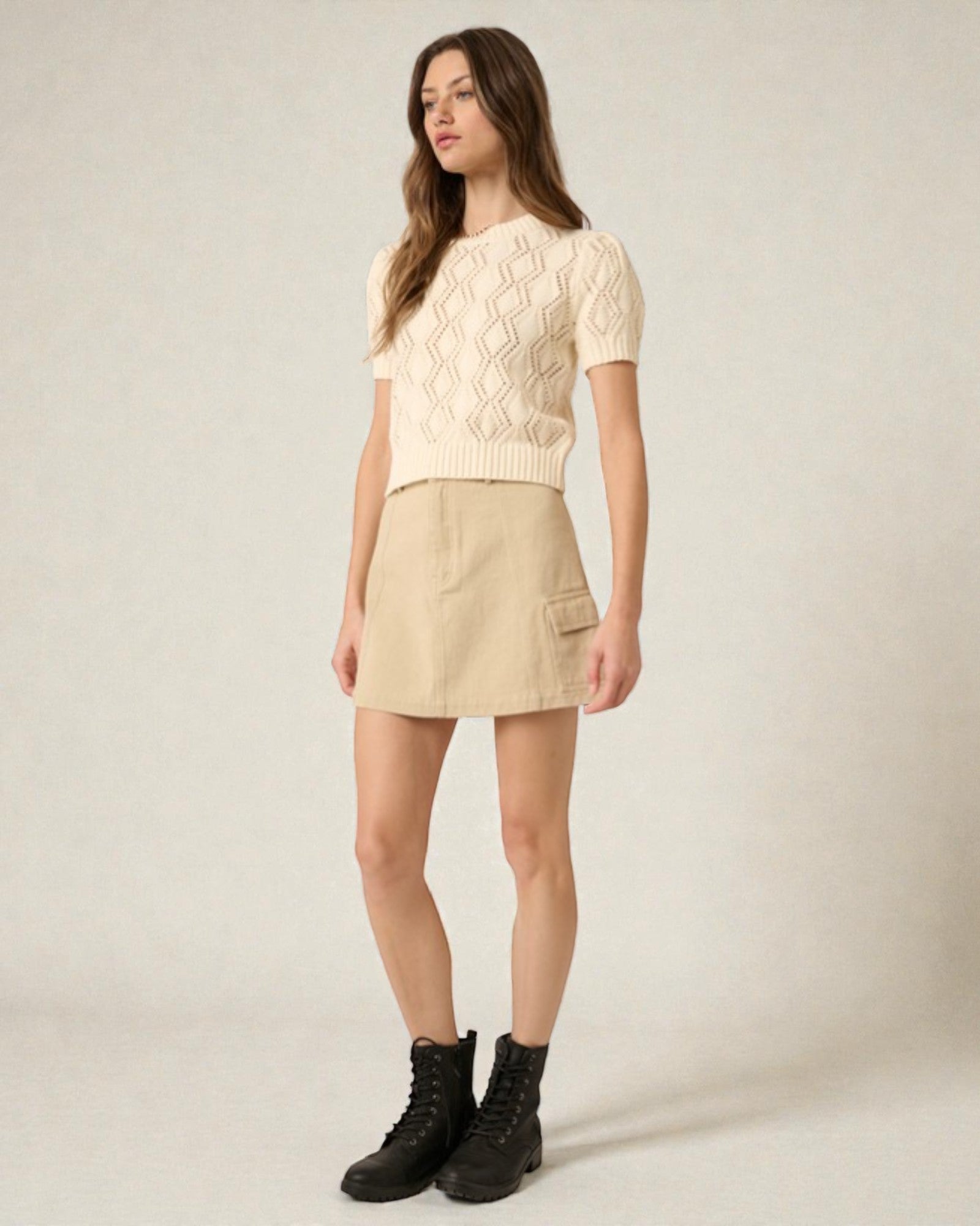 Woman wearing a beige cable knit sweater and skirt with black boots on a plain background