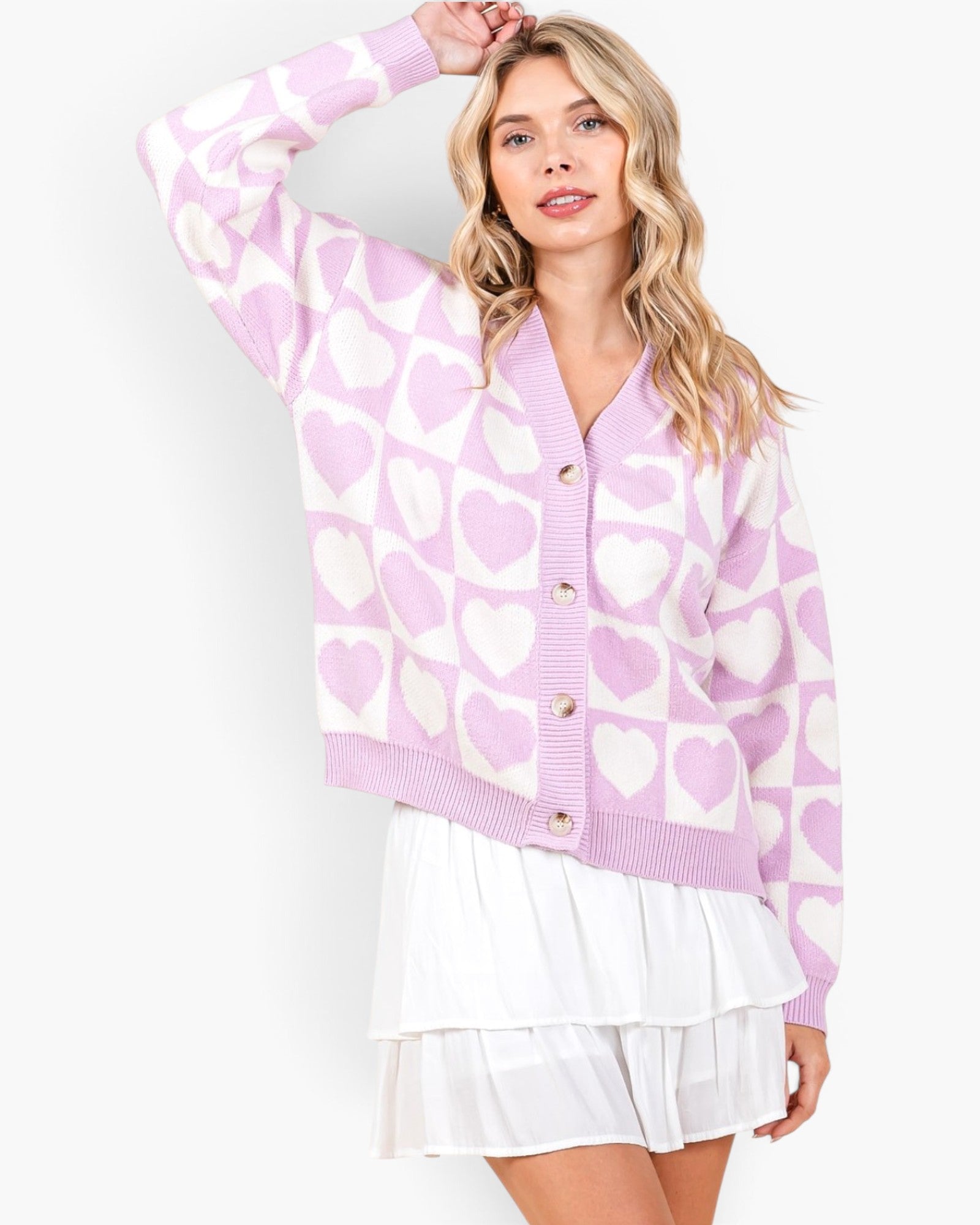 A woman wearing a pink and white knit cardigan with a heart pattern, paired with a white tiered skirt, against a plain light gray studio background.