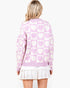 A woman viewed from the back wearing a light pink and white knit cardigan with a heart pattern, layered over a white ruffled mini skirt, against a plain white background.