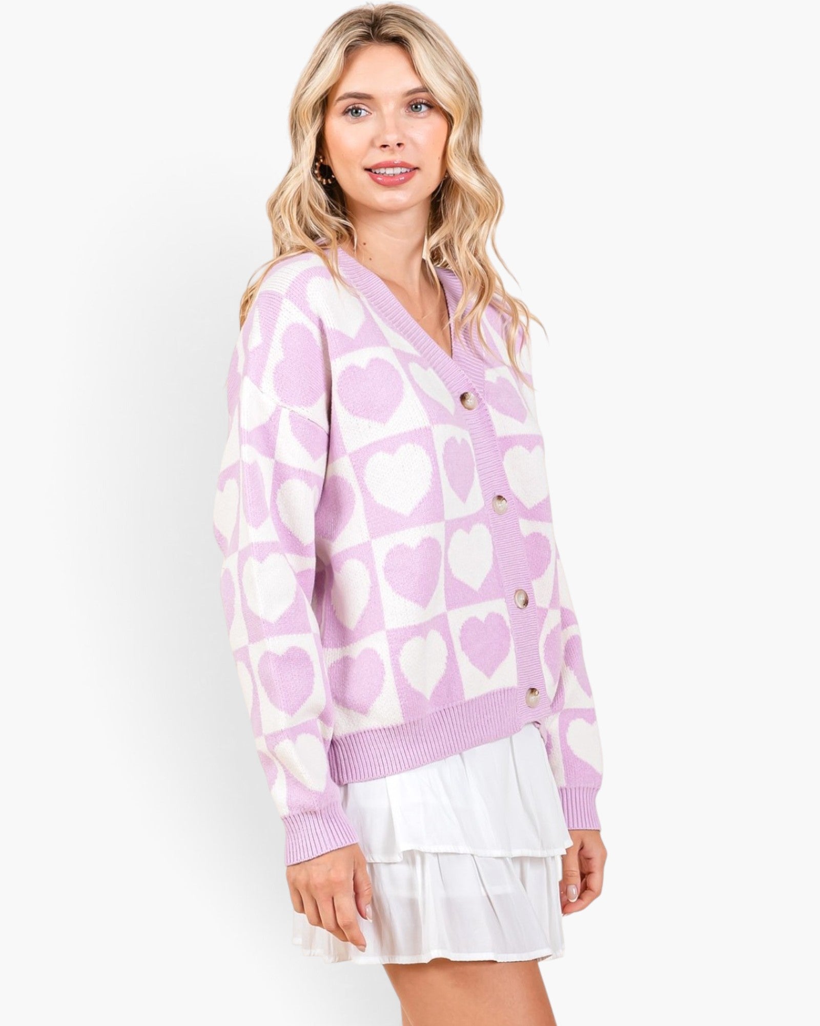 A woman wearing a lavender and white heart-patterned knit cardigan over a white ruffled mini skirt, photographed against a clean white studio background.