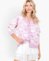 A woman wearing a pink and white heart-patterned knit cardigan with gold buttons, paired with a white tiered skirt, against a plain white studio background.
