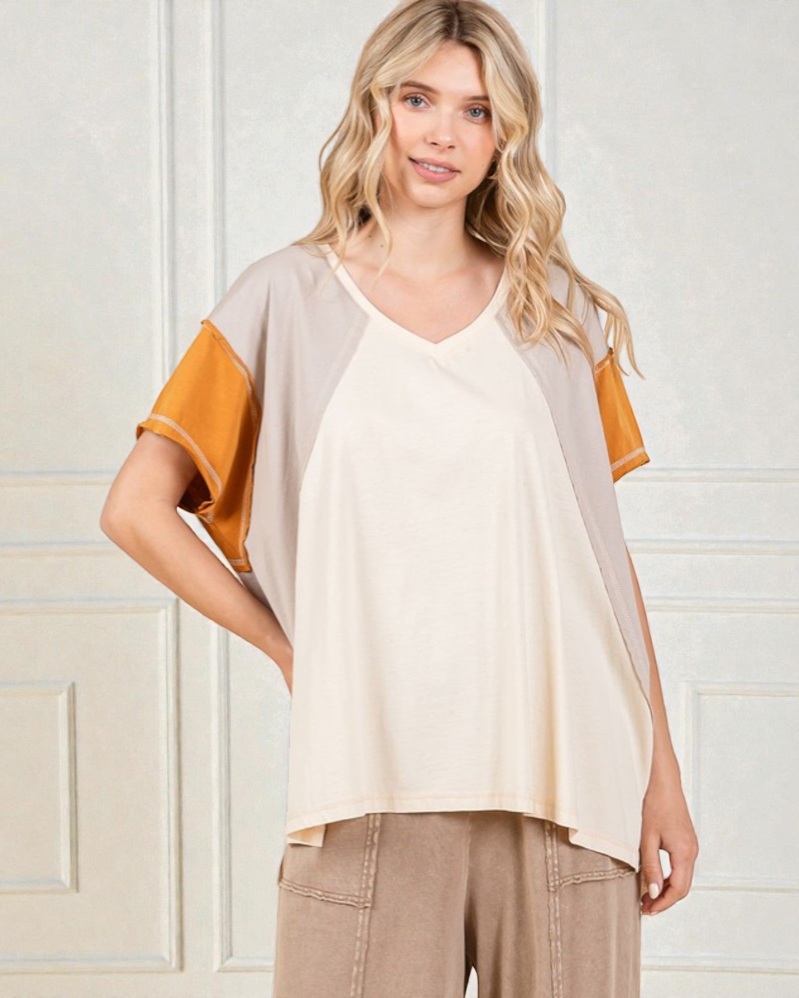 Woman wearing a beige and orange top with beige pants against a white wall.
