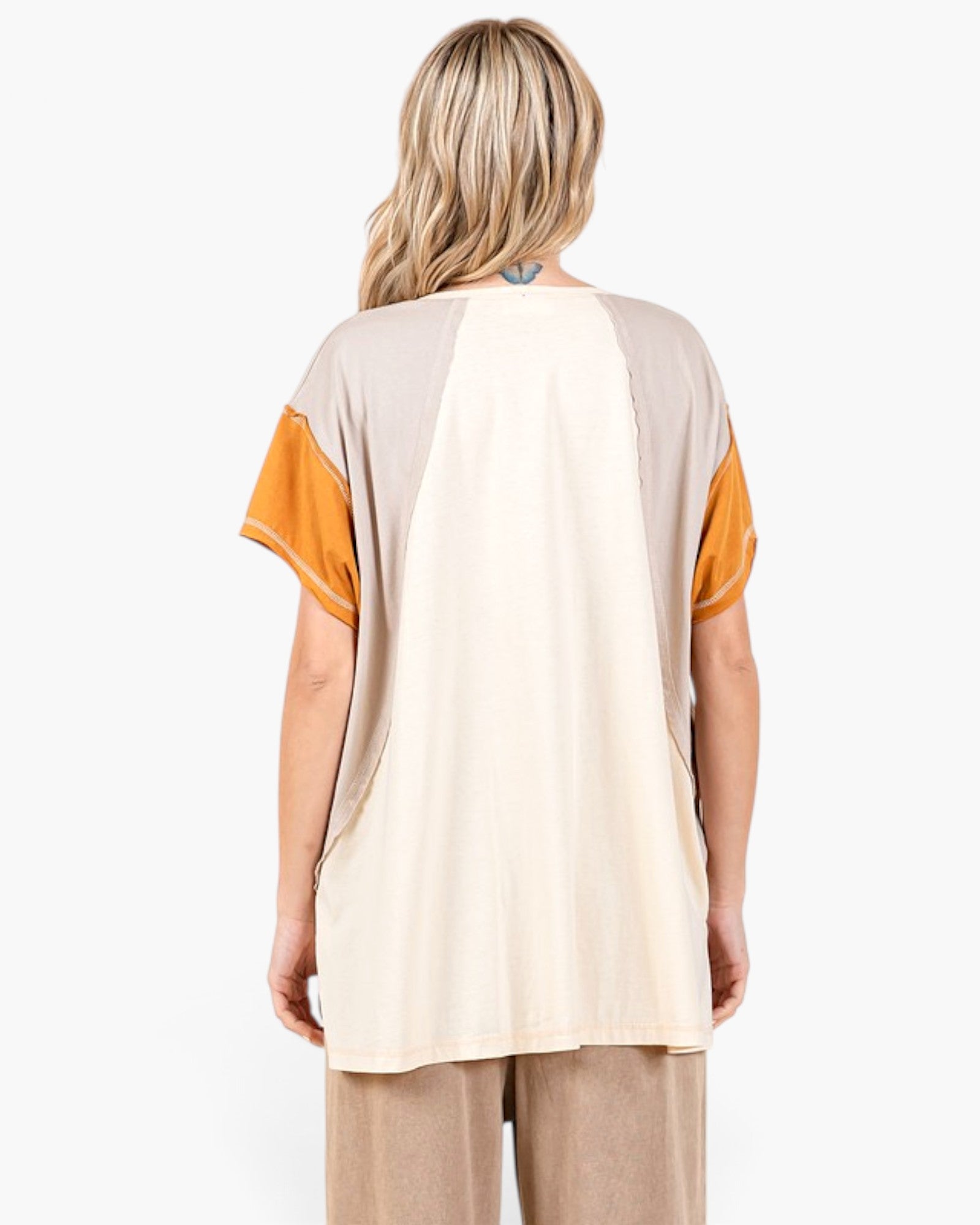 A woman viewed from the back wearing a color-block short-sleeve top with a large cream center panel, taupe shoulder panels, and mustard yellow sleeves, paired with beige pants against a plain white background.