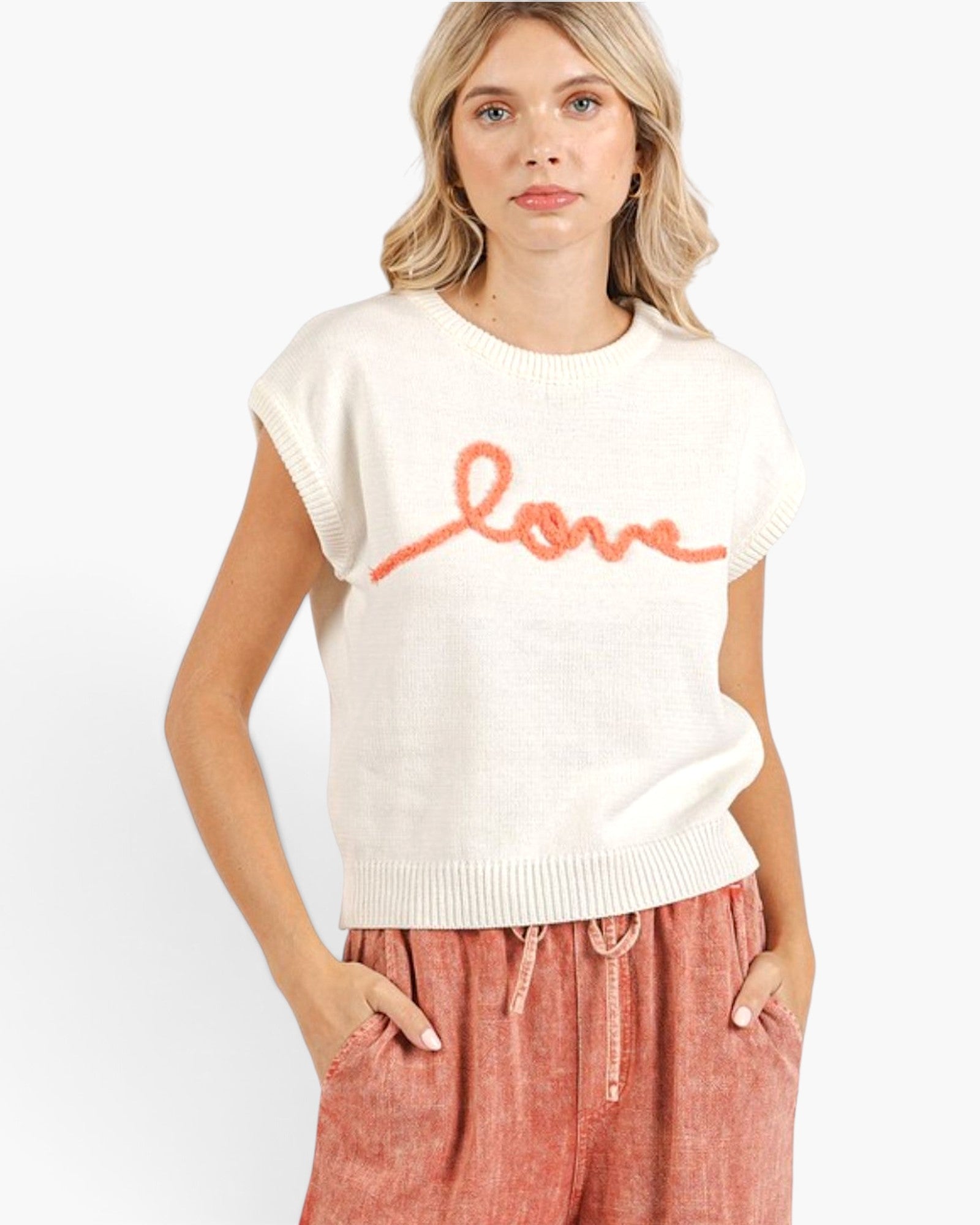 A woman wearing a cream-colored sleeveless knit sweater with the word 'love' written across the chest in orange, paired with dusty rose drawstring pants, against a plain white background.