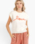 A woman wearing a cream-colored sleeveless knit sweater with the word 'love' written across the chest in orange, paired with dusty rose drawstring pants, against a plain white background.