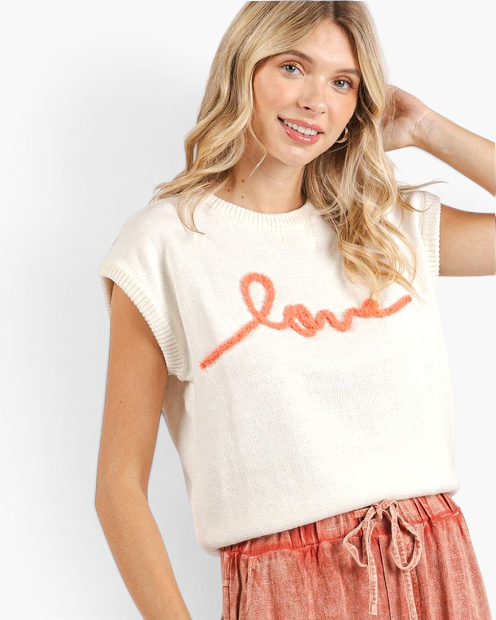 A woman wearing a cream-colored knit sweater with a textured orange 'love' script across the chest, paired with pink drawstring pants, against a plain white studio background.