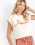 A woman wearing a cream-colored knit sweater with a textured orange 'love' script across the chest, paired with pink drawstring pants, against a plain white studio background.