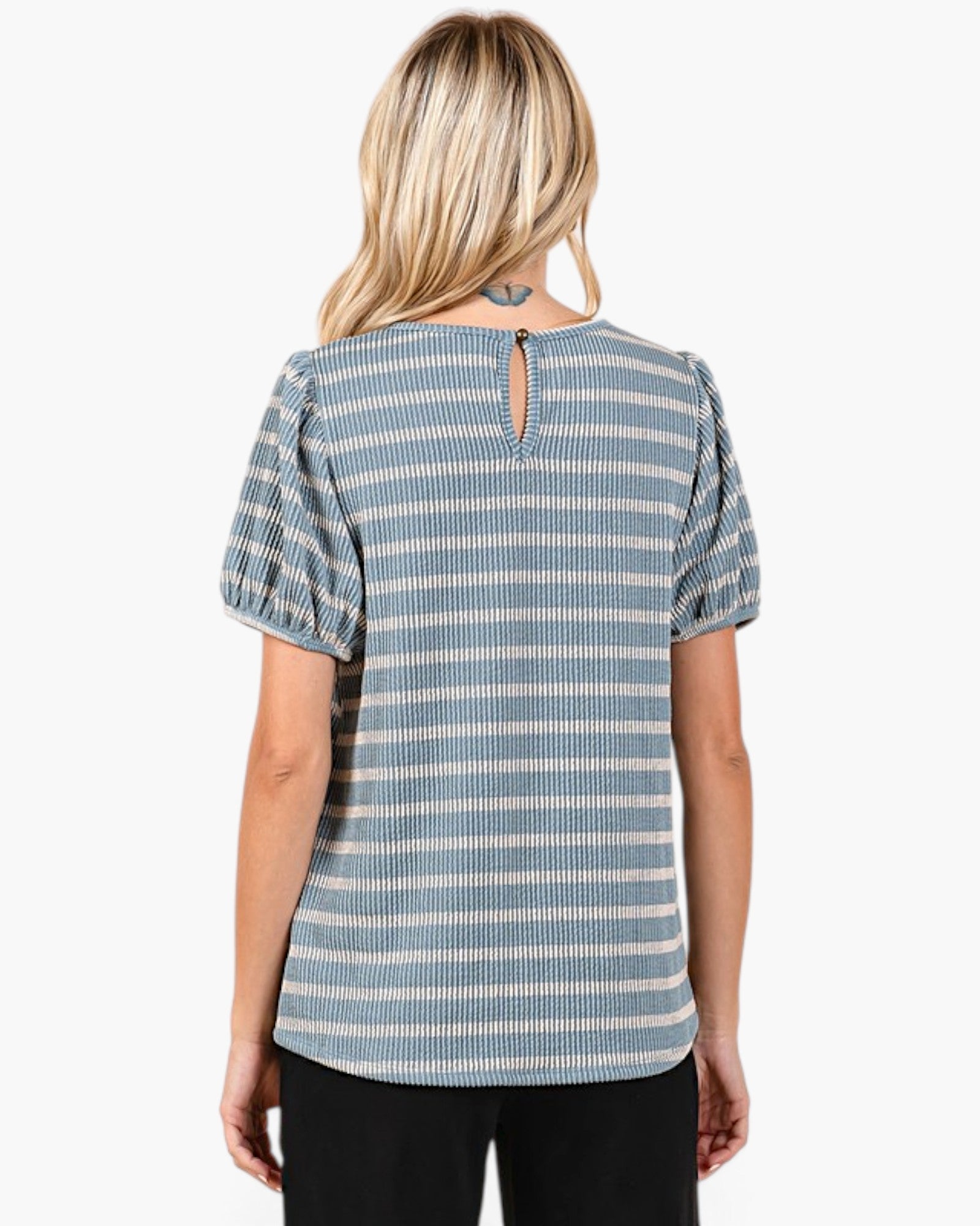 A woman viewed from the back wearing a short-sleeve top with vertical blue and off-white stripes, a small keyhole cutout at the back neckline, and puff sleeves. She is also wearing black pants against a plain light gray studio background.