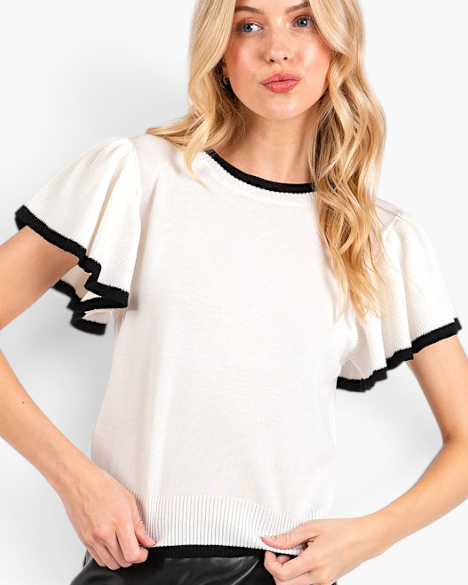 A blonde woman wearing a cream-colored short-sleeve knit top with black ruffled trim on the sleeves and neckline, paired with black bottoms, against a plain white background.