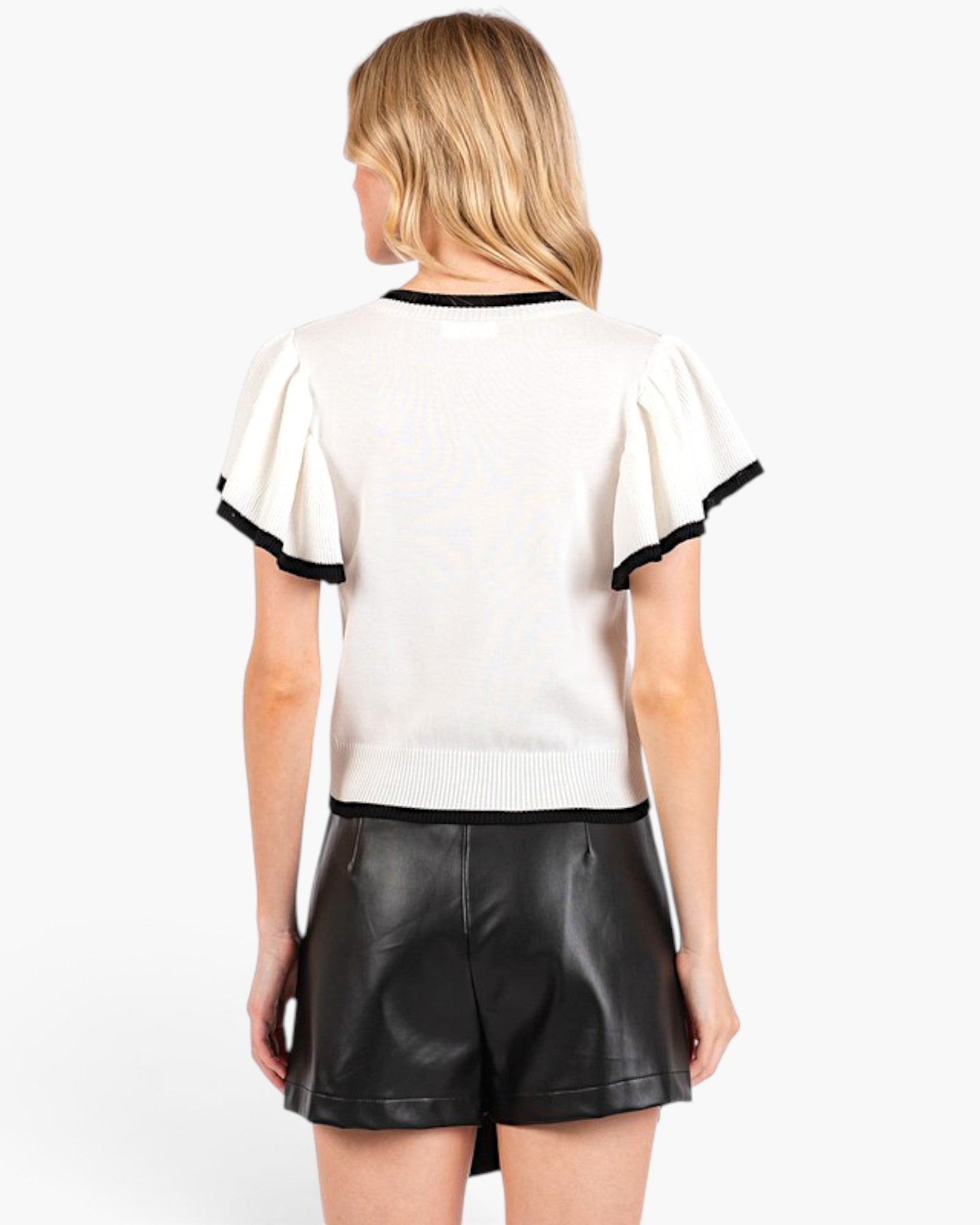 A woman shown from the back wearing a cream knit short-sleeve top with black trim and black faux-leather shorts against a plain white studio background.