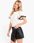 A blonde woman modeling a white short-sleeve top with black ruffle trim on the sleeves and collar, paired with black faux-leather shorts, against a plain white studio background.
