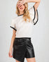 Woman wearing a white top with black trim and black leather shorts on a light gray background