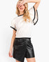 A woman modeling a white short-sleeve knit top with black trim and black faux leather wrap shorts against a plain white background.