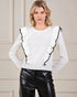 Woman wearing a white sweater with black ruffled details and black leather pants against a neutral background