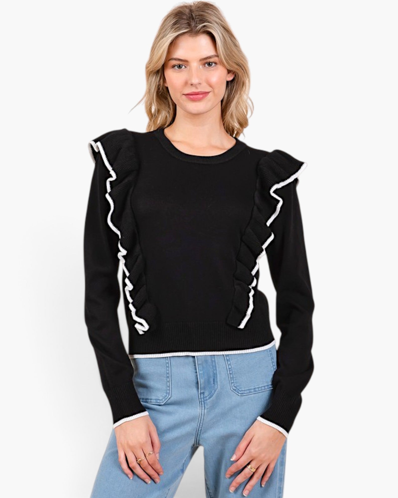 A woman modeling a black long-sleeve knit top with white ruffle trim along the shoulders and cuffs, paired with light blue jeans, against a plain white studio background.