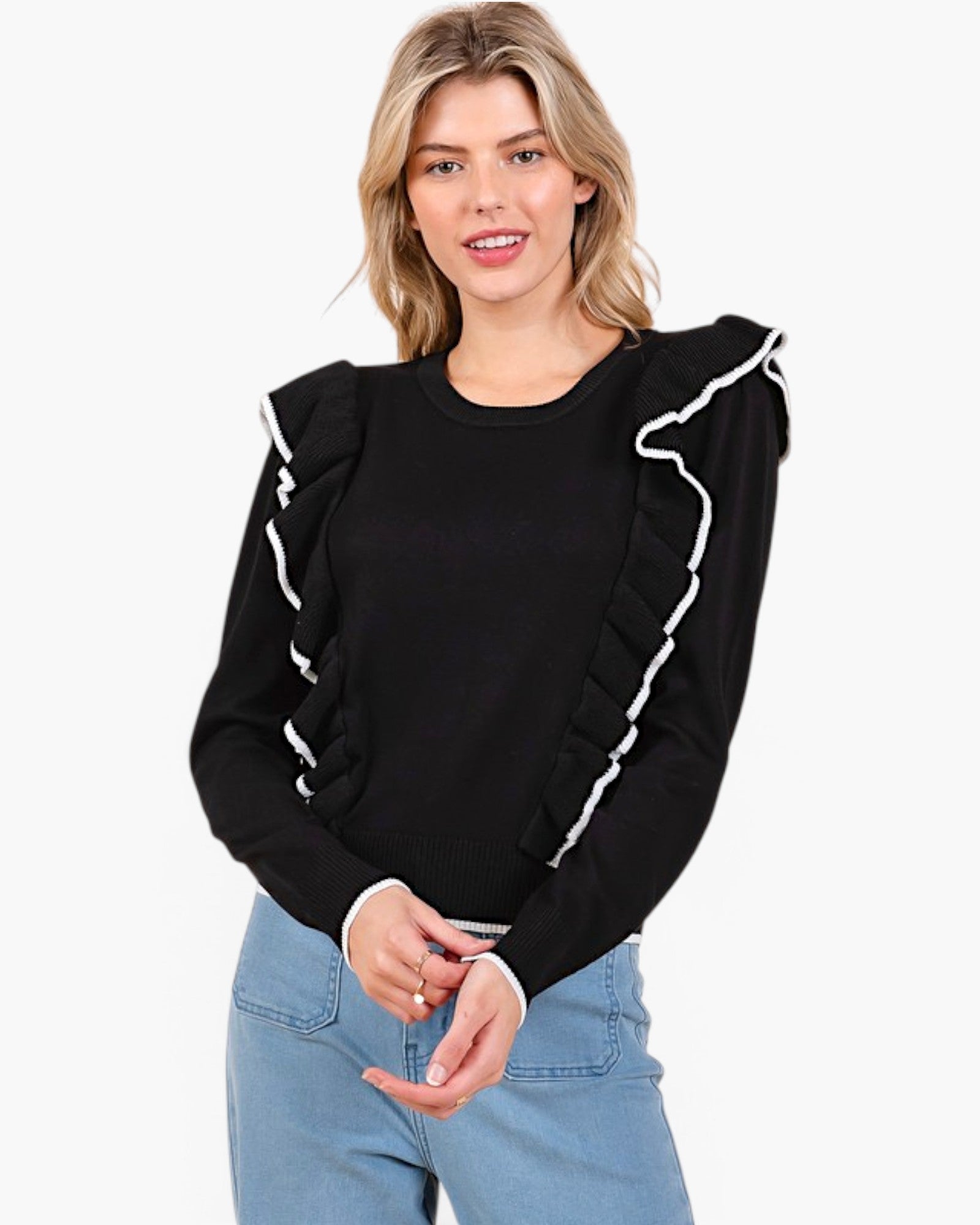 A woman wearing a black long-sleeve knit top with white ruffle trim along the shoulders and sleeves, paired with light blue jeans, against a plain white background.